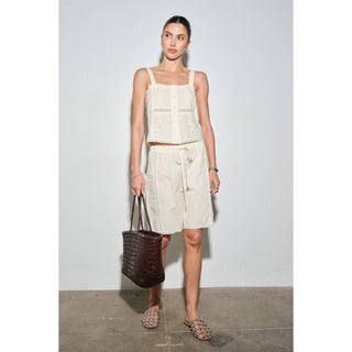 A woman models a matching two-piece linen set consisting of a cream sleeveless crop top with lace trim and matching drawstring shorts, holding a dark brown woven tote bag.
