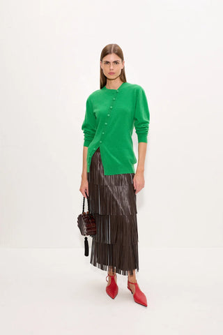 A model stands against a plain white studio background wearing a bright green button-front cardigan, a dark brown pleated midi skirt, and red pointed heels, while holding a small dark handbag.