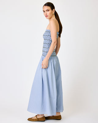 A woman modeling a strapless light blue maxi dress with a smocked bodice and a subtle gingham pattern on the skirt, paired with brown flat shoes against a plain white studio background.