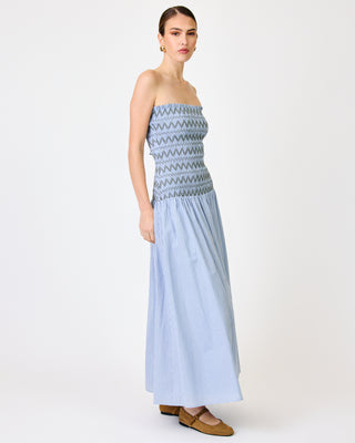 A woman modeling a strapless maxi dress with a light blue and white vertically striped skirt and a smocked bodice featuring a gray and blue geometric pattern, set against a plain light gray studio background.