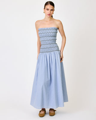 A woman modeling a strapless light blue and white gingham maxi dress with a smocked bodice featuring a geometric pattern, set against a plain white studio background.