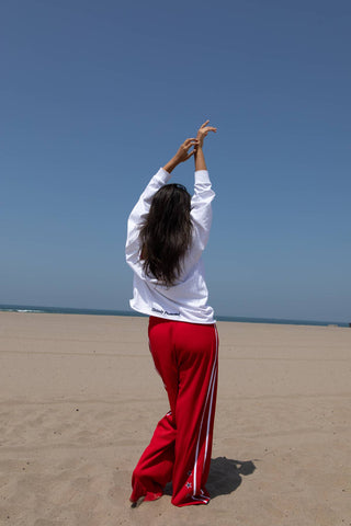 A woman standing on a sandy beach with her back to the camera, wearing a white long-sleeve top and bright red track pants with white side stripes, with her arms raised above her head.