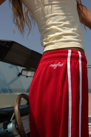 A close-up lifestyle shot of a person wearing red athletic shorts with white side stripes and a white script logo that reads 'mifas' on the hip, standing near a boat.