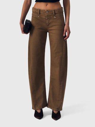 A model wearing a pair of wide-leg brown jeans, holding a black clutch, against a light gray studio background.