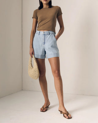 Woman wearing a brown top and light blue shorts, holding a straw bag against a neutral background