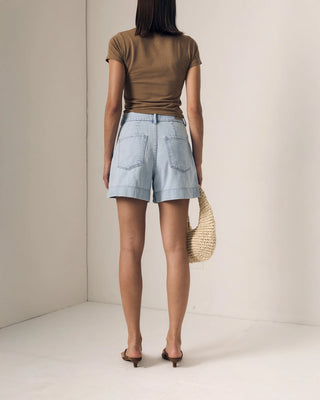 Person wearing a brown top and light blue shorts holding a woven bag against a plain background
