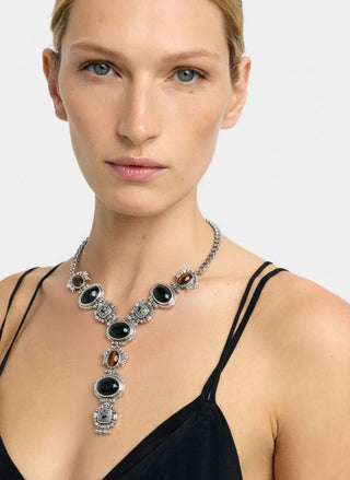 A close-up of a model wearing a chunky silver statement necklace with black oval stones and amber accents, paired with a black spaghetti-strap top against a light gray background.
