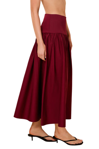 A woman wearing a high-waisted, pleated maxi skirt in deep burgundy, paired with black thong sandals, shown from the waist down against a plain white background.