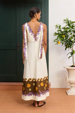 Woman in a long, patterned dress standing in a room with a plant and door.