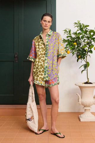 Woman in a colorful patterned outfit holding a bag, standing next to a plant indoors.
