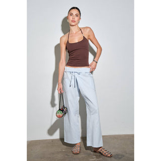 Woman wearing a brown tank top and light blue pants against a white wall.