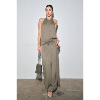 A woman models a sleeveless, high-neck, floor-length draped dress in a muted taupe-brown color, holding a small woven handbag against a plain studio backdrop.