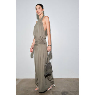 A woman modeling a sleeveless, halter-neck, floor-length draped dress in taupe. She is holding a textured dark gray handbag and wearing white pointed-toe heels, standing against a plain light gray studio wall.