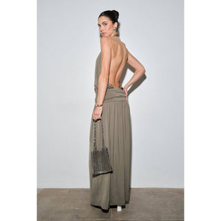 A woman stands with her back to the camera wearing a draped, open-back maxi dress in a muted taupe-brown color. She is holding a small woven handbag against a plain light gray studio wall.