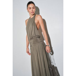 A woman modeling a sleeveless, halter-neck maxi dress in a muted olive green color, holding a silver chain handbag against a plain light gray studio background.