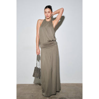 A woman models a sleeveless, high-neck, floor-length taupe dress with a ruched waist, holding a textured silver handbag against a light gray studio background.