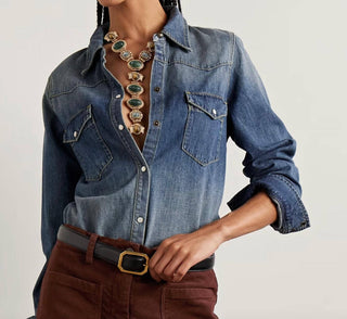 A person wearing a blue denim western-style shirt with chest pockets, paired with brown pants and a chunky statement necklace, photographed against a plain light background.