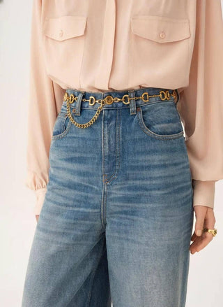 A close-up of a person wearing high-waisted blue jeans with a decorative gold chain belt and a blush pink blouse against a light neutral background.