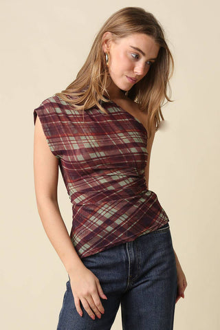 A woman wearing a one-shoulder wrap top with a burgundy, green, and beige plaid pattern, paired with dark blue jeans, against a solid beige background.