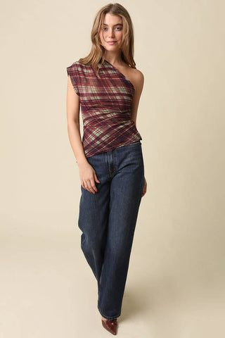 A woman modeling a one-shoulder wrap top with a burgundy, green, and beige plaid pattern, paired with dark blue wide-leg jeans, against a solid beige background.