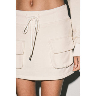 Close-up of a model wearing a cream-colored mini skirt with a drawstring waist and large front flap pockets.