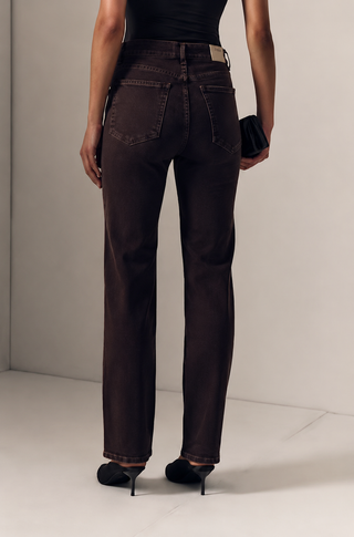 A model shown from the back wearing dark brown straight-leg jeans, a black top, and black high heels, holding a black clutch against a minimalist light gray studio background.