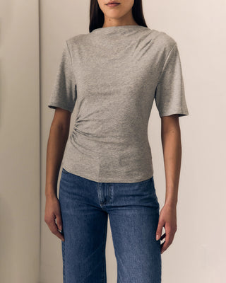 A woman wearing a light heather gray short-sleeve t-shirt with a ruched side detail and blue jeans, shown from the back against a neutral beige background.