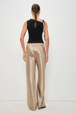 A woman shown from the back wearing a black sleeveless top and shiny beige wide-leg trousers with a small repeating dark dot pattern, against a plain light gray studio background.