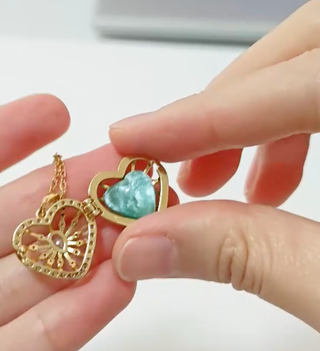 Gold heart-shaped pendant with a turquoise stone held between fingers on a light background