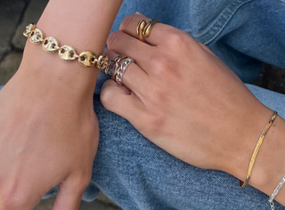 A close-up lifestyle shot of a person's hands and wrist wearing multiple gold jewelry pieces, including a chunky interlocking link bracelet on the wrist, several stacked rings on the fingers, and a delicate gold chain bracelet on the other wrist, set against blue denim jeans.