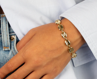 A gold-toned chain bracelet with oval links worn on a person's wrist, shown against a white shirt and blue denim background.
