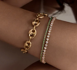 Two bracelets worn on a wrist: a gold chain bracelet with oval links and a second bracelet featuring green and clear stones in a heart pattern.