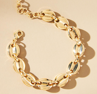 A gold chain bracelet with polished oval links, arranged in a loose circle on a warm beige background with soft shadows.