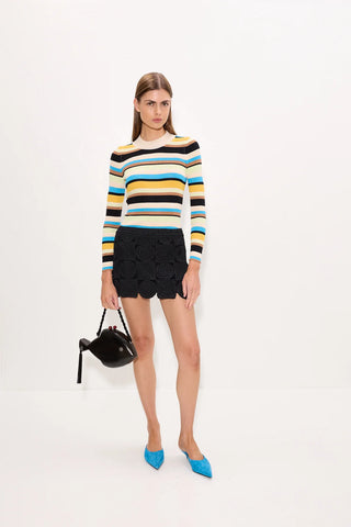 A model stands against a plain white studio background wearing a long-sleeve striped knit top with black textured shorts and bright blue pointed shoes, while holding a small black handbag.