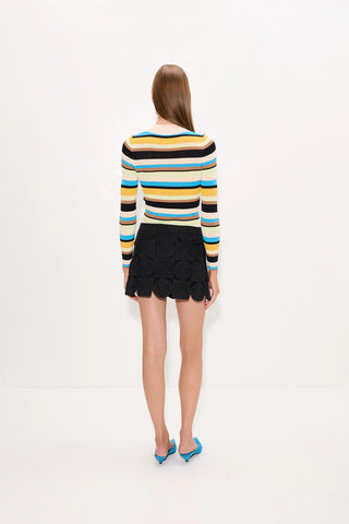 A model viewed from behind wearing a colorful horizontally striped long-sleeve knit top with black textured mini skirt and blue pointed-toe heels, set against a plain white studio background.