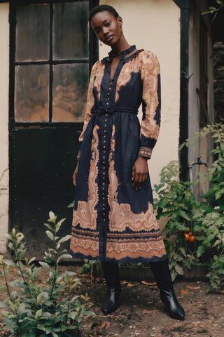 A woman stands outdoors in front of a rustic cream-colored wall and dark door, wearing a long-sleeved midi dress with a navy blue base and a beige paisley pattern. The dress features a button-up front, a matching waist tie, and a high collar.
