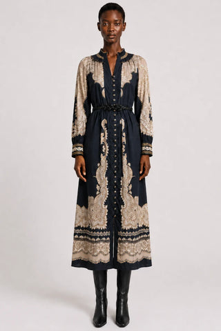 A model stands against a light gray studio background wearing a long-sleeved midi dress with a navy blue base and beige paisley embroidery, paired with black ankle boots.