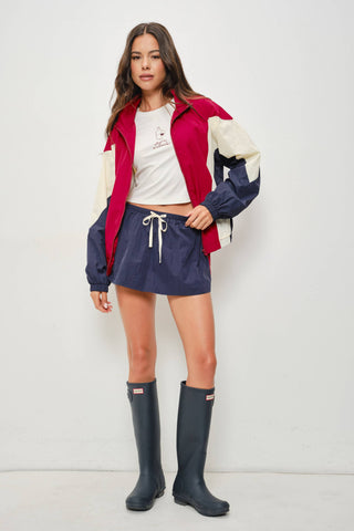 A woman modeling a colorblock windbreaker jacket with navy blue, burgundy red, and cream panels, paired with a white crop top, navy blue drawstring skirt, and dark navy rain boots against a plain white studio background.