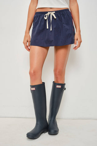 A model wearing a navy blue corduroy mini skirt with a cream drawstring, paired with navy blue rain boots. The background is a clean, off-white studio setting.