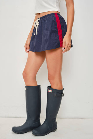 A model from the waist down wearing navy blue shorts with a red side stripe and white drawstring, paired with dark navy rain boots against a plain white studio background.