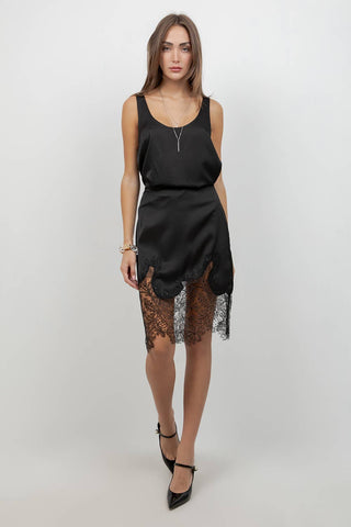 A woman modeling a black satin slip dress with a sheer black lace hem, paired with black pointed-toe heels, against a plain light gray studio background.