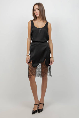 A woman models a black satin slip dress with a sheer black lace overlay at the hem, paired with black pointed-toe heels, against a plain light gray studio background.