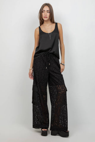 A model stands against a light gray studio background wearing a black satin sleeveless top and black lace wide-leg cargo pants with side flap pockets.