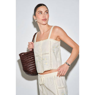 A woman modeling a cream-colored embroidered crop top and matching drawstring pants, holding a dark brown woven tote bag against a plain light gray background.