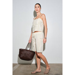 A woman models a matching two-piece crochet set consisting of a cream camisole top and shorts, holding a dark brown woven tote bag against a plain light gray studio background.