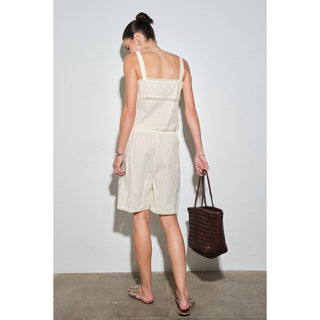 A woman is shown from behind wearing a light cream-colored sleeveless romper with a textured lace-like pattern on the back. She is holding a dark brown woven handbag and wearing flat sandals, standing against a plain light gray wall.