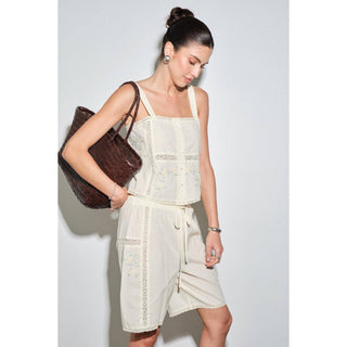 A woman modeling a cream-colored two-piece linen set with delicate floral embroidery and lace trim, carrying a brown woven tote bag against a light gray studio background.