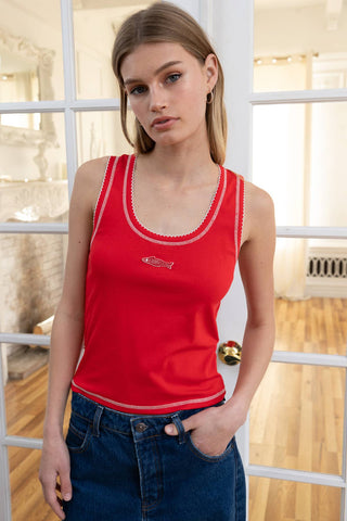 Woman wearing a red tank top with a fish logo in a room with wooden floor and white walls.