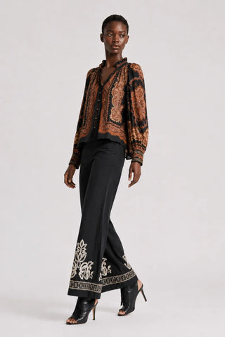 A model is shown from the head to toe wearing a black and rust-orange paisley patterned button-up shirt, black wide-leg trousers with white floral embroidery at the hem, and black high-heeled ankle boots, against a light gray studio background.